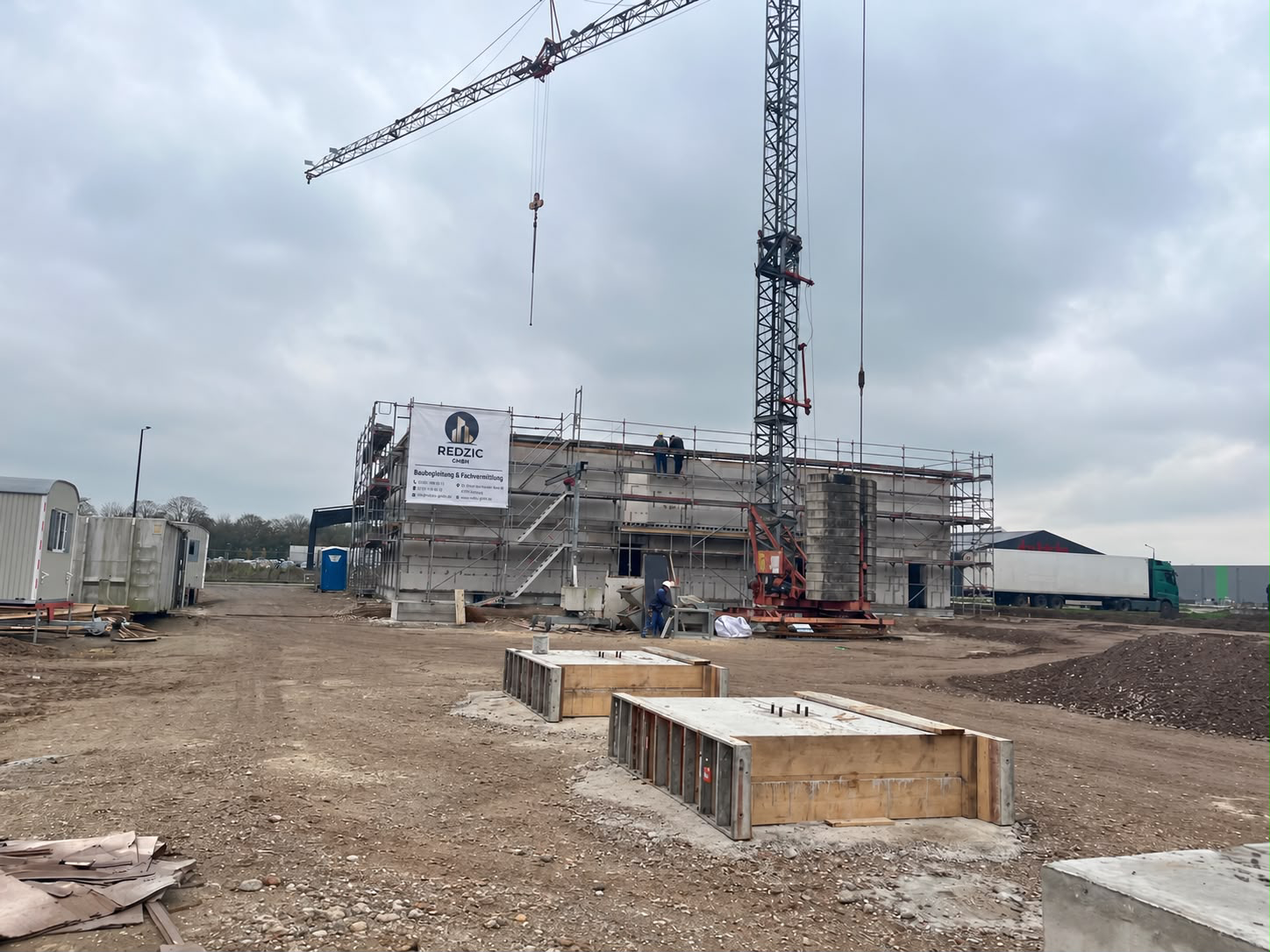 Baustelle des Gewerbegebäudes mit Turmdrehkran, Fundamentschalungen im Vordergrund und Lagerhalle im Hintergrund